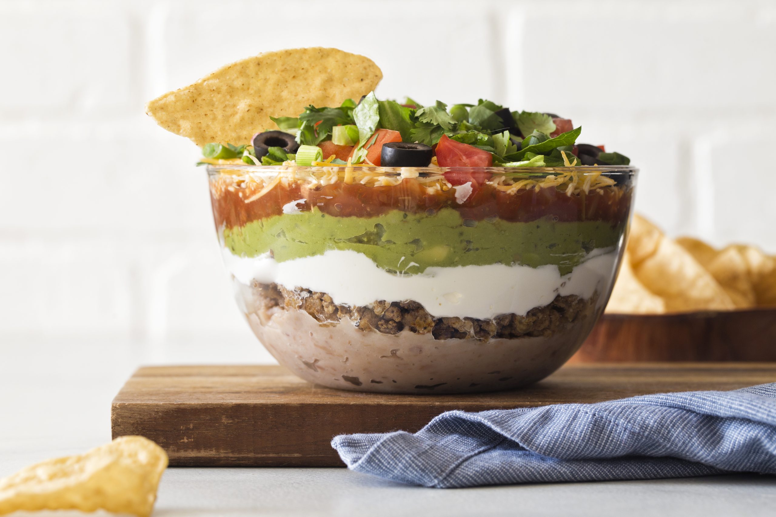 7 Layer Taco Dip, THE tailgating snack featuring layers of seasoned ground veal, creamy refried beans, sour cream, fresh guacamole and more!